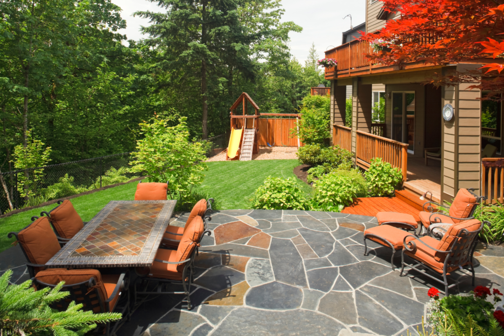 A natural stone patio with a patio furniture dining table and chairs and a kids play area in the backyard and surrounded by bushes.