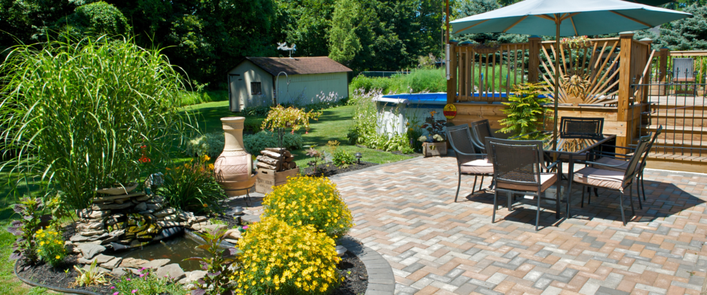 A full stone patio with a dining set and bushes surrounding it and a pergola cover.