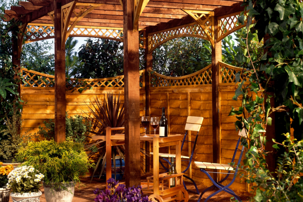 A patio with a surrounding privacy fence and a pergola over the patio providing shade and bushes planters on the patio.