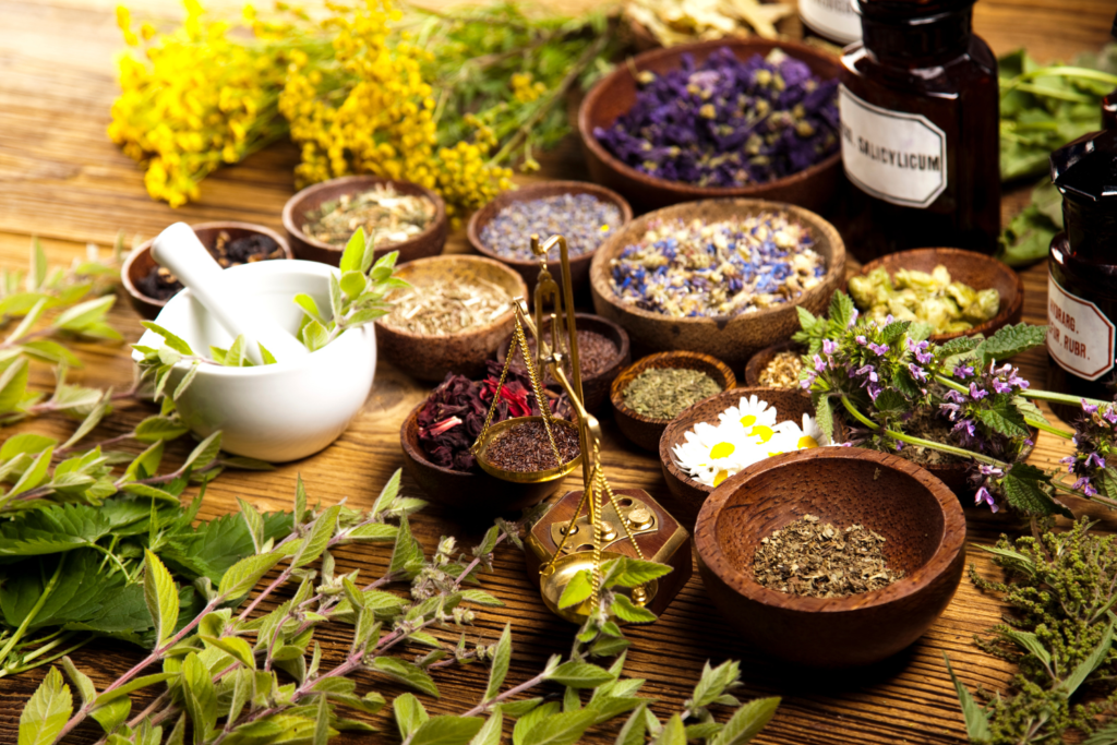 A mortar and pestle on a wood table with various herbs and a scale.