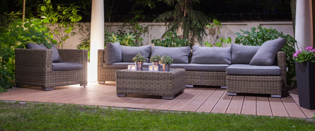 A stone patio with gray wicker furniture and grey cushions and pillows and a pergola over it and bushes surrounding it.