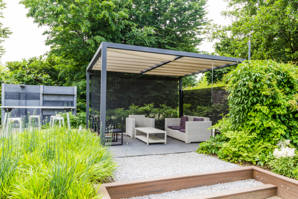 A cement patio with a gray pergola over it and blue furniture with cushions and pillows and bushes surrounding it.