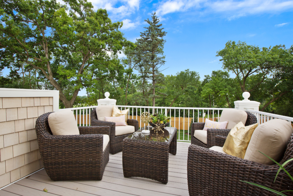 A patio with brown wicker furniture on a deck overlooking trees in the backyard and decorative pillow on the chairs.