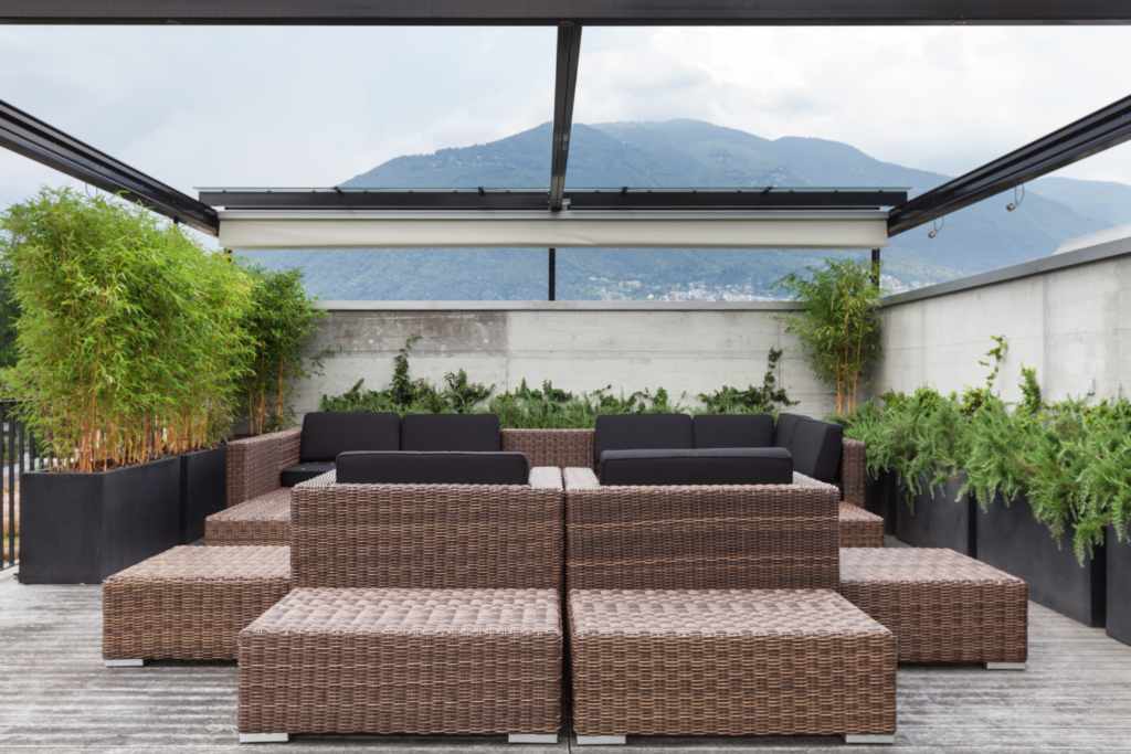 A patio with brown wicker furniture and a glass pergola roof over the top and bushes surrounding the patio.