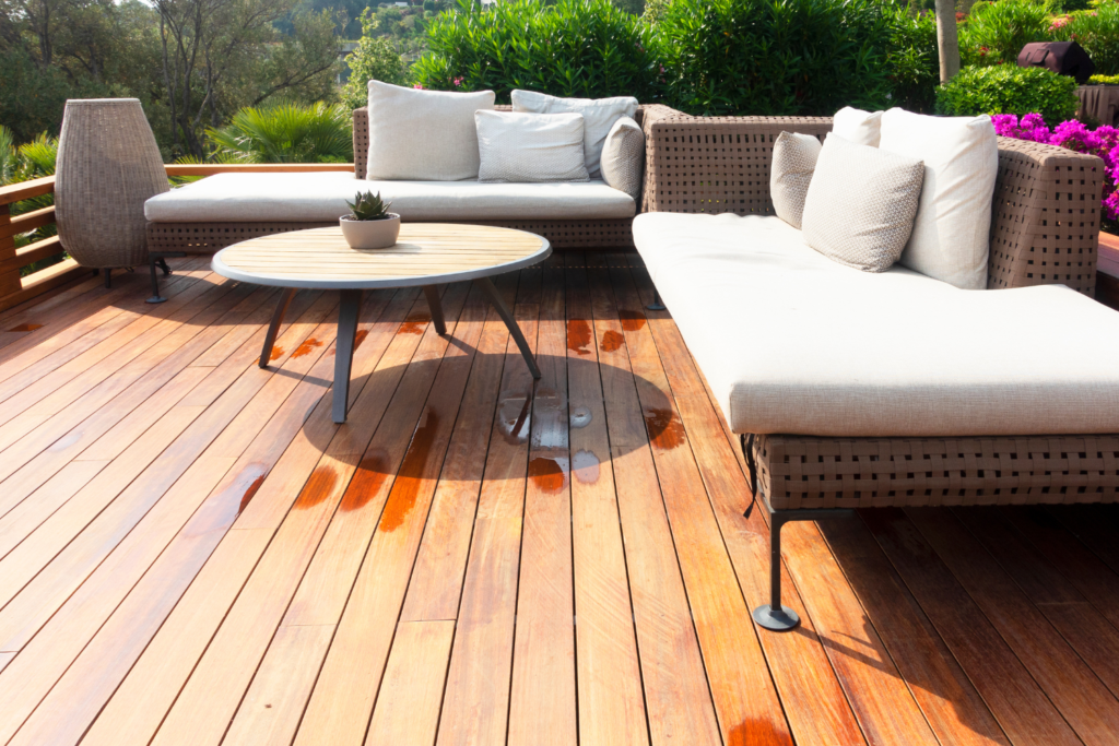 An L shaped patio with tan wicker furniture and white cushions and pillows with a small round coffee table and trees in the background.