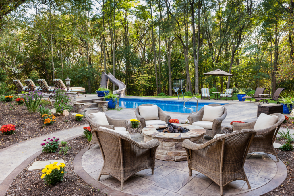 A stone patio with tan wicker furniture and a firepit sitting next to a pool and trees in the backyard.