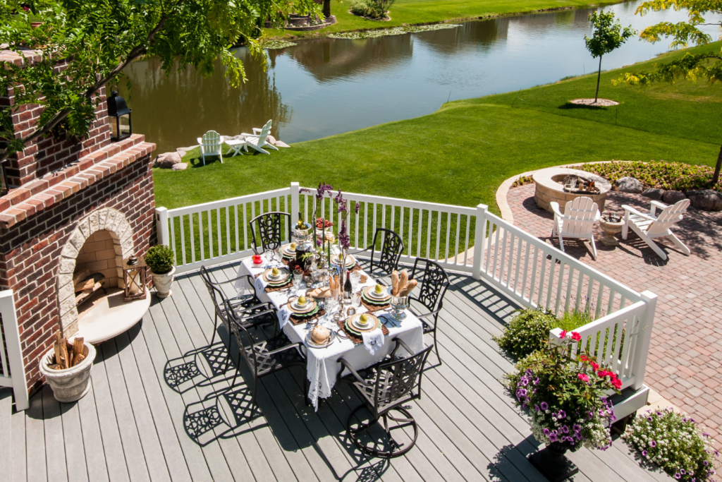 A multi -level patio deck with a dining area and a sitting area and a stone fireplace along side of the dining table with a grass backyard and lake.