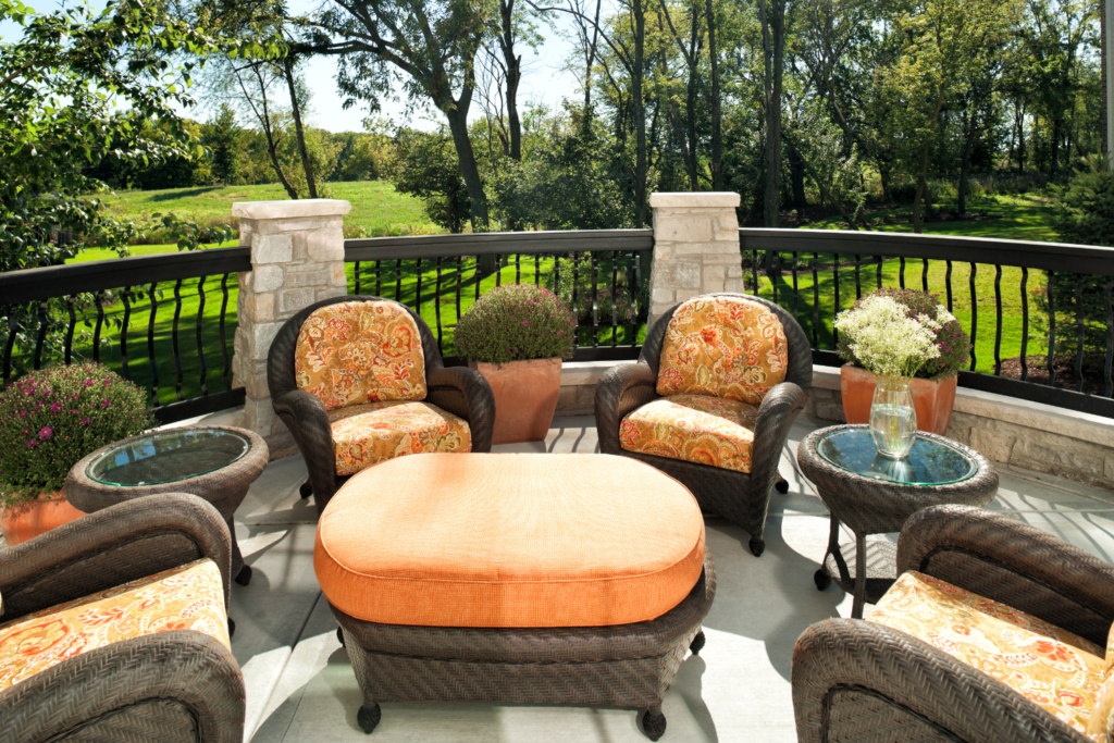 A patio with brown wicker furniture and orange cushions and pillows and a ottoman and overlooking the backyard full of trees.