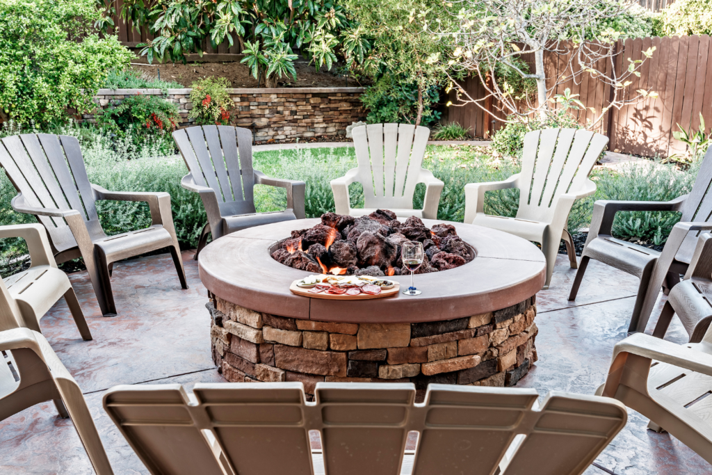 A stone patio with tan furniture in a round layout design with a stone firepit in the middle and trees and a privacy fence in the backyard.