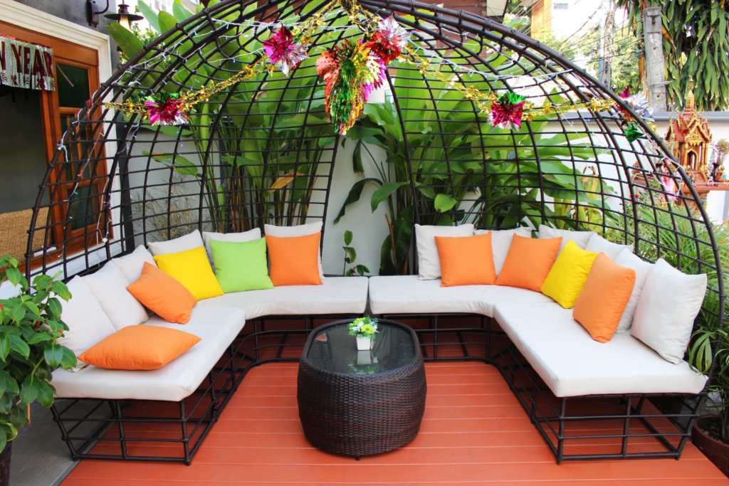 A U shaped patio furniture set with black wicker and white cushions and yellow and orange pillows and a tall bushy plant behind it.