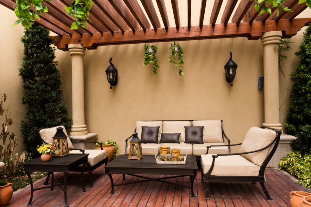 A patio with a pergola over it and brown furniture with tan pillows with sconces on the house walls.