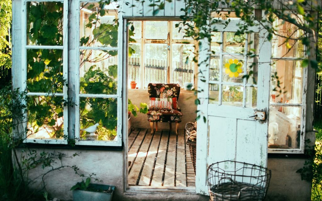 A white interior view of a she shed with vines hanging down, a chair on the inside and glass windows all around.